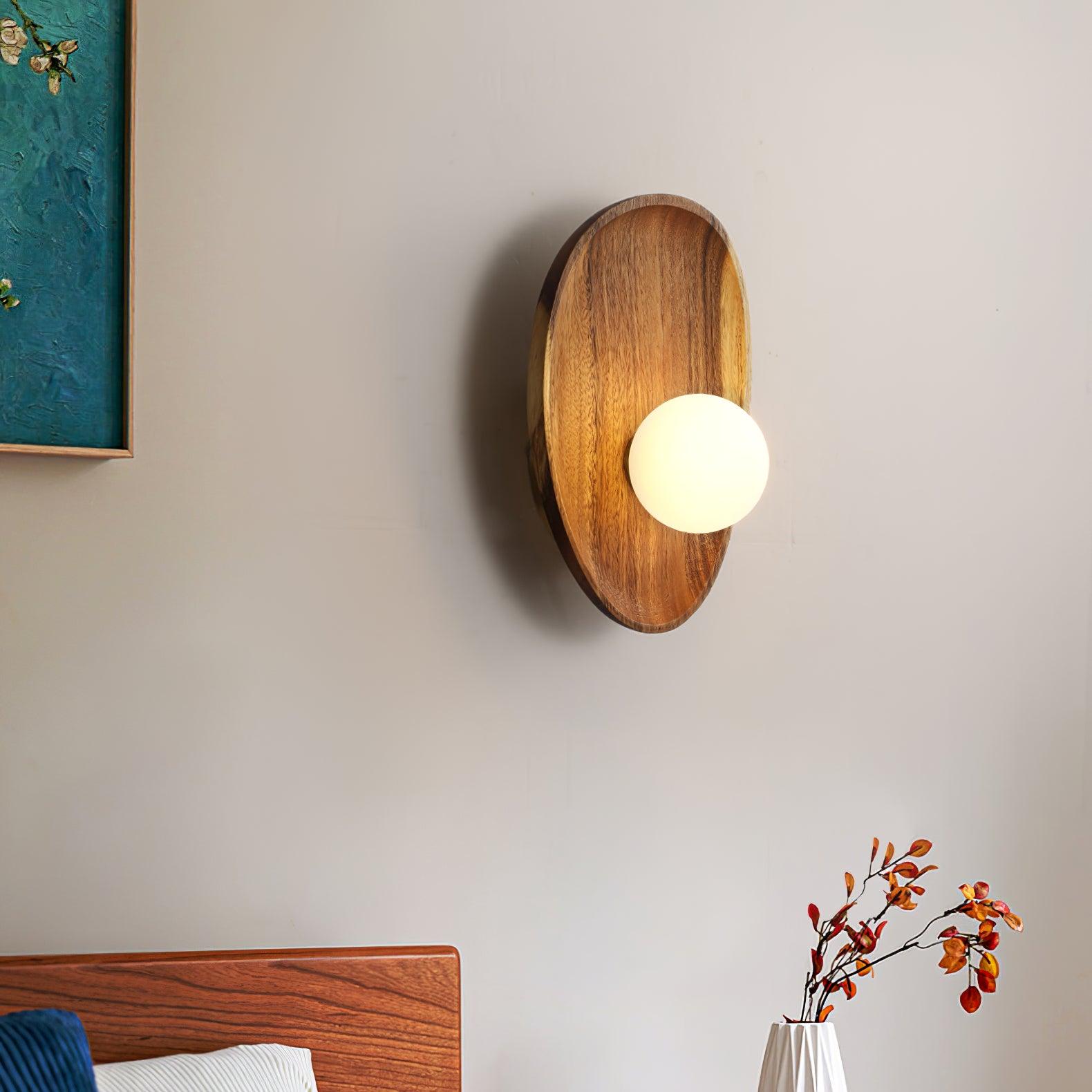 Wall lamp with a shaped walnut-coloured wooden backplate and a white glass globe lit beside a framed picture and a bed.