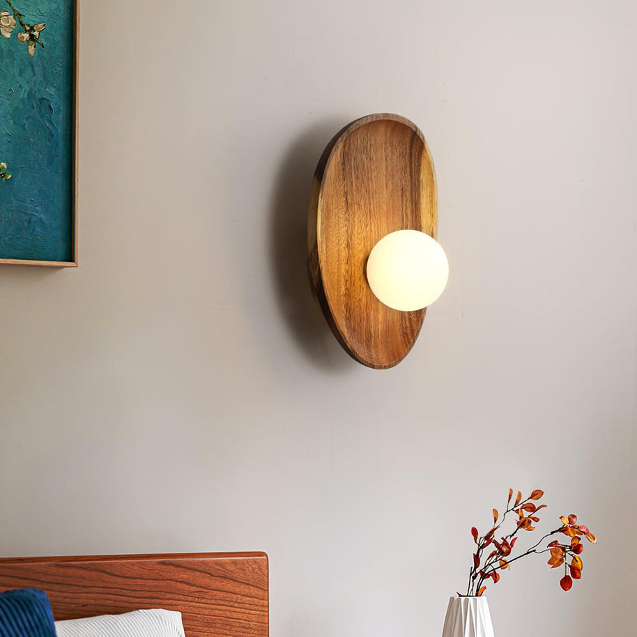 Wall lamp with a shaped walnut-coloured wooden backplate and a white glass globe lit beside a framed picture and a bed.
