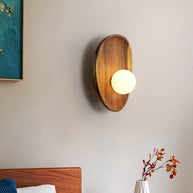 Wall lamp with a shaped walnut-coloured wooden backplate and a white glass globe lit beside a framed picture and a bed.