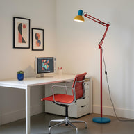 Adjustable floor lamp with a red arm, yellow shade and round blue base beside a white desk and a red office chair.