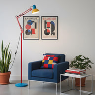 Adjustable floor lamp with a red arm, yellow shade and round blue base beside a blue armchair below two framed prints.