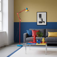 Adjustable floor lamp with a red arm, yellow shade and round blue base in a living room with a grey sofa and a two-tone wall.
