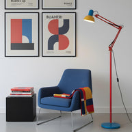 Adjustable floor lamp with a red arm, yellow shade and round blue base beside a blue armchair below framed posters.