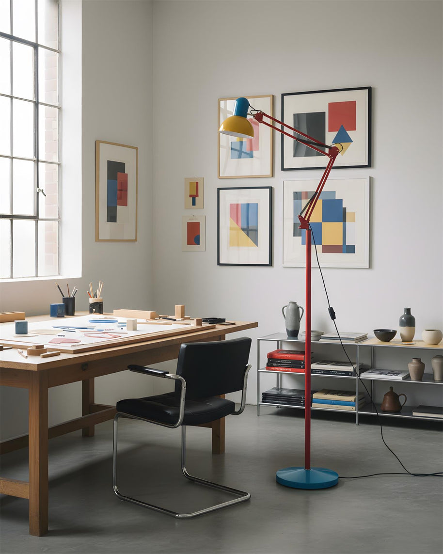 Adjustable floor lamp with a red arm, yellow shade and round blue base in a studio beside a large wooden work table.