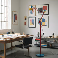 Adjustable floor lamp with a red arm, yellow shade and round blue base in a studio beside a large wooden work table.