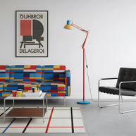 Adjustable floor lamp with a red arm, yellow shade and round blue base in a living room with a colour-block sofa and a poster.