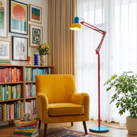 Adjustable floor lamp with a red arm, yellow shade and round blue base lit beside a yellow armchair and a bookshelf.