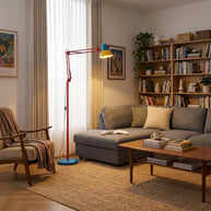 Adjustable floor lamp with a red arm, yellow shade and round blue base in a living room with a grey sofa and a bookcase.