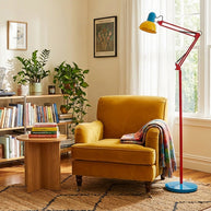 Adjustable floor lamp with a red arm, yellow shade and round blue base beside a mustard armchair at a sunlit window.