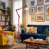 Adjustable floor lamp with a red arm, yellow shade and round blue base in a living room with a blue sofa, bookshelf and framed prints.