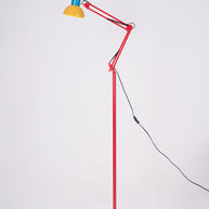 Adjustable floor lamp with a red arm, yellow shade and round blue base with the arm angled forward, on a plain background.