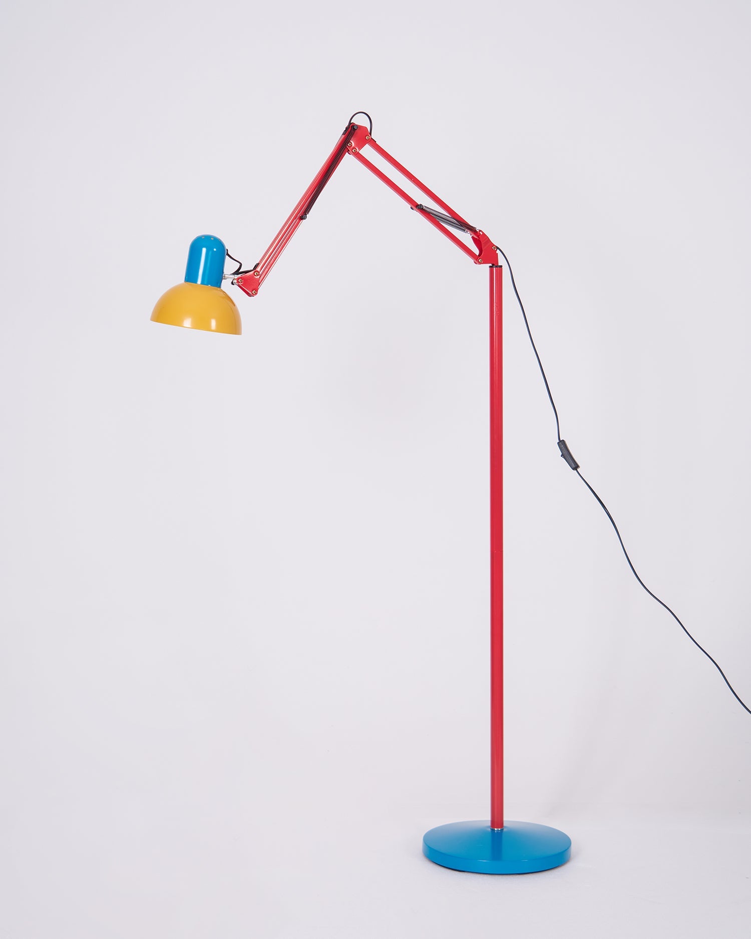 Adjustable floor lamp with a red arm, yellow shade and round blue base with the arm extended low, on a plain background.