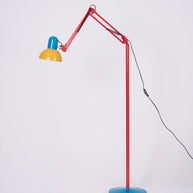 Adjustable floor lamp with a red arm, yellow shade and round blue base with the arm extended low, on a plain background.