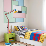 Adjustable floor lamp with a red arm, yellow shade and round blue base beside a child's bed in a room with a pastel panelled wall.