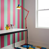 Adjustable floor lamp with a red arm, yellow shade and round blue base beside a desk against a boldly striped wall.