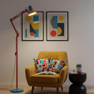 Adjustable floor lamp with a red arm, yellow shade and round blue base beside a mustard armchair on a round striped rug.