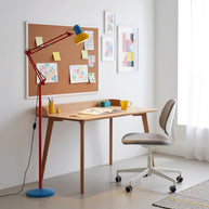 Adjustable floor lamp with a red arm, yellow shade and round blue base beside a wooden desk and a pinboard.