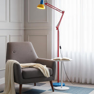 Adjustable floor lamp with a red arm, yellow shade and round blue base beside a grey armchair with a throw.