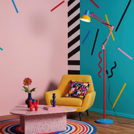 Adjustable floor lamp with a red arm, yellow shade and round blue base in a living area with a yellow armchair against pink and teal walls.