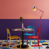 Adjustable floor lamp with a red arm, yellow shade and round blue base beside a dining table in a room with pink and purple walls.