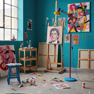 Adjustable floor lamp with a red arm, yellow shade and round blue base in an artist's studio beside an easel.