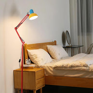 Adjustable floor lamp with a red arm, yellow shade and round blue base lit beside a wooden bed.