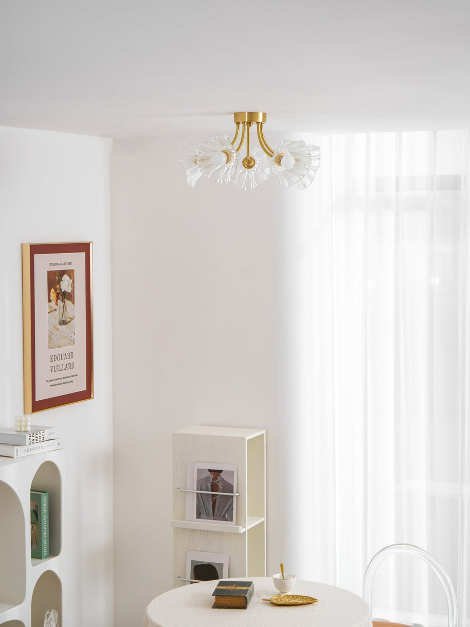 Flush-mount ceiling lamp with curved brass arms holding clear ribbed flower-shaped glass shades lit on a ceiling above a table and shelving.