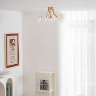 Flush-mount ceiling lamp with curved brass arms holding clear ribbed flower-shaped glass shades lit on a ceiling above a table and shelving.