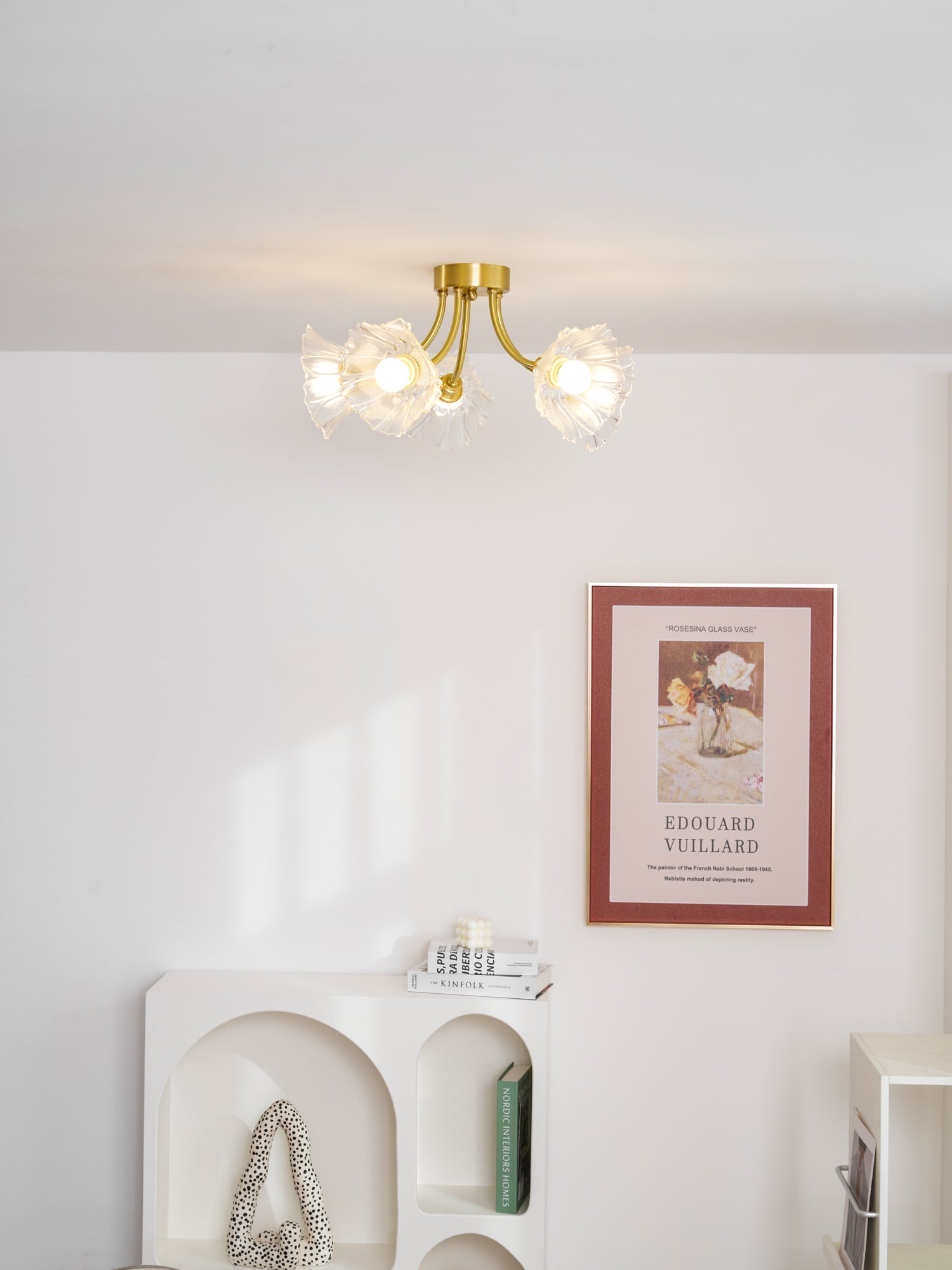 Flush-mount ceiling lamp with curved brass arms holding clear ribbed flower-shaped glass shades lit on a ceiling above an alcove shelf.