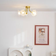 Flush-mount ceiling lamp with curved brass arms holding clear ribbed flower-shaped glass shades lit on a ceiling above an alcove shelf.