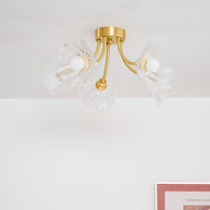 Flush-mount ceiling lamp with curved brass arms holding clear ribbed flower-shaped glass shades, unlit, seen against a pale ceiling.