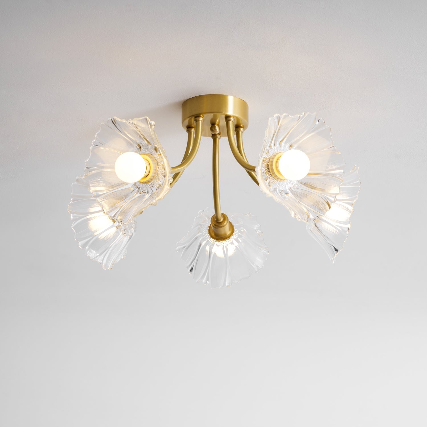 Flush-mount ceiling lamp with curved brass arms holding clear ribbed flower-shaped glass shades, lit, seen close against a pale ceiling.