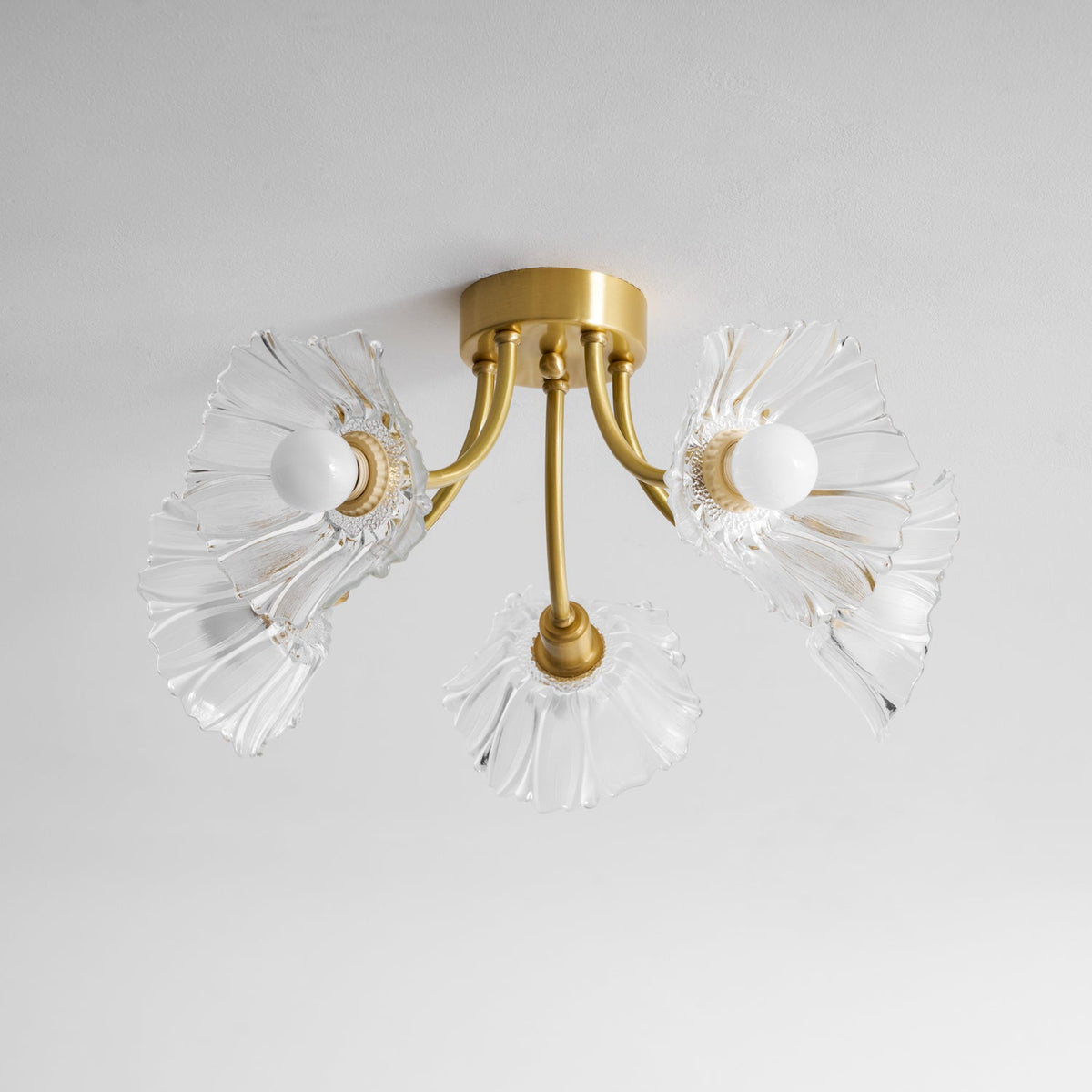 Flush-mount ceiling lamp with curved brass arms holding clear ribbed flower-shaped glass shades, lit, seen from below and to one side.