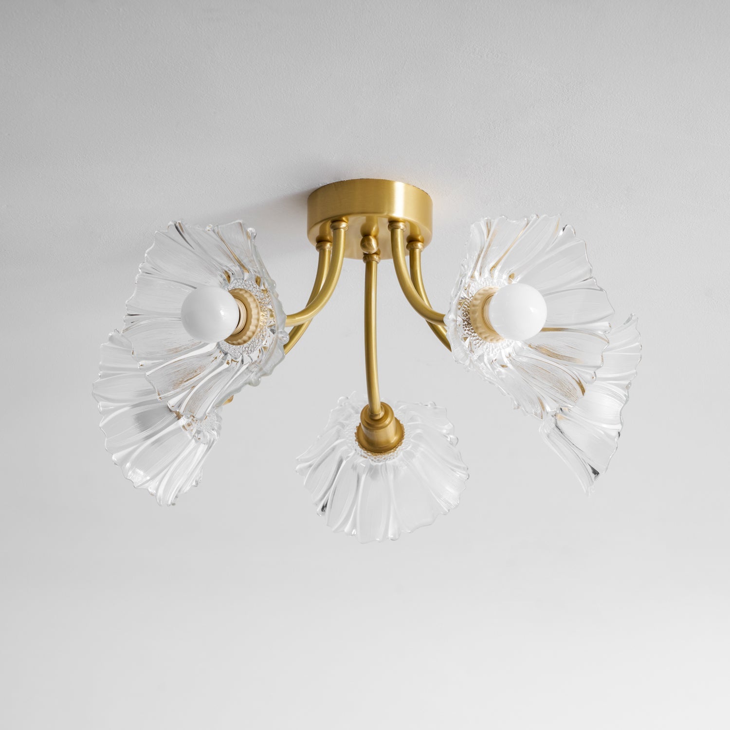 Flush-mount ceiling lamp with curved brass arms holding clear ribbed flower-shaped glass shades, lit, seen from below and to one side.