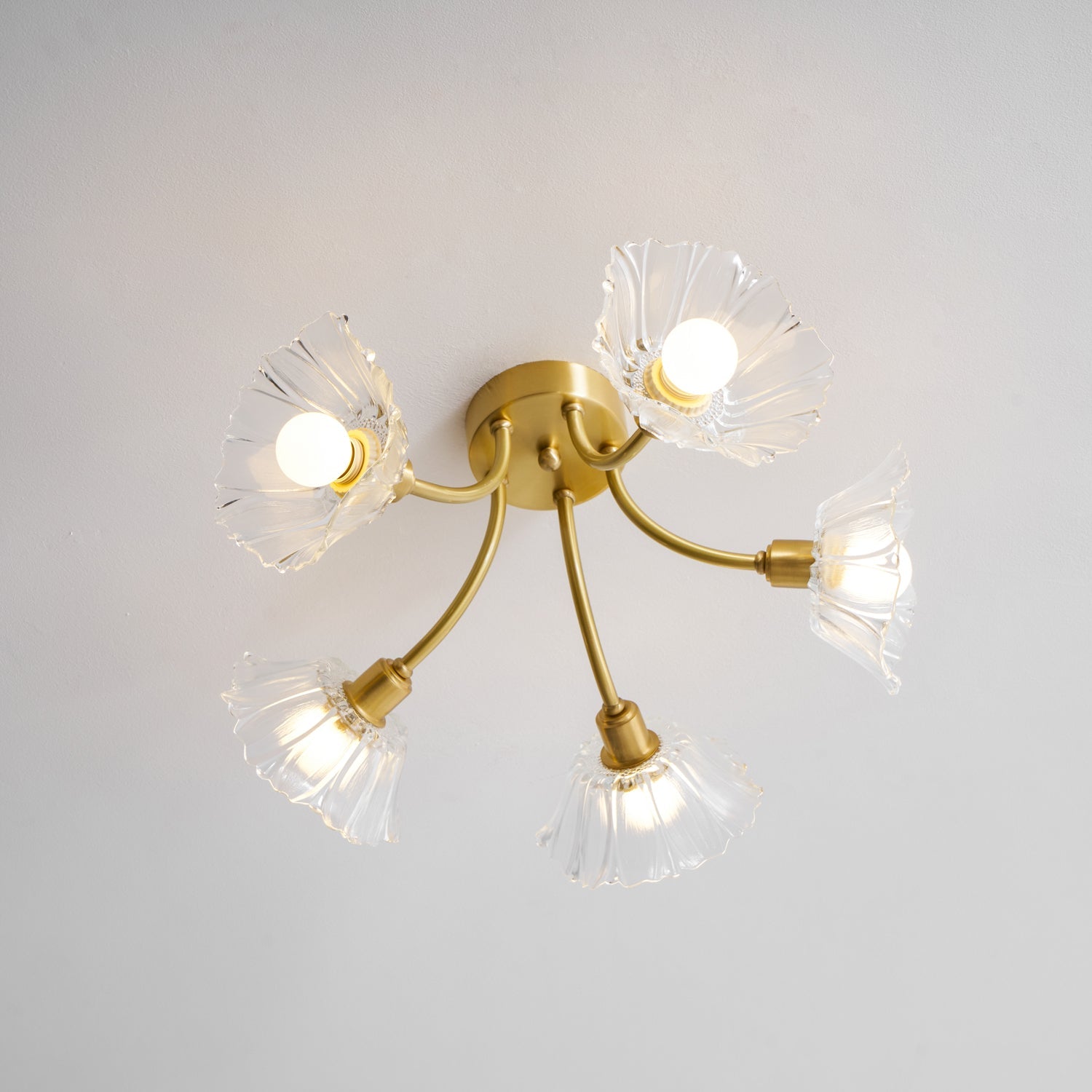 Flush-mount ceiling lamp with curved brass arms holding clear ribbed flower-shaped glass shades, lit, seen from below with the arms spread wide.