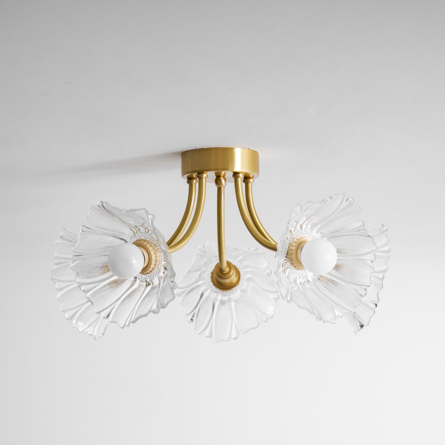 Flush-mount ceiling lamp with curved brass arms holding clear ribbed flower-shaped glass shades, lit, seen against a ceiling edge.