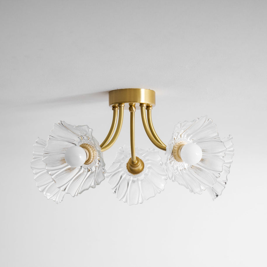 Flush-mount ceiling lamp with curved brass arms holding clear ribbed flower-shaped glass shades, lit, seen against a ceiling edge.