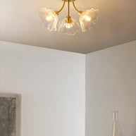 Flush-mount ceiling lamp with curved brass arms holding clear ribbed flower-shaped glass shades lit on a ceiling in a corner of a room.