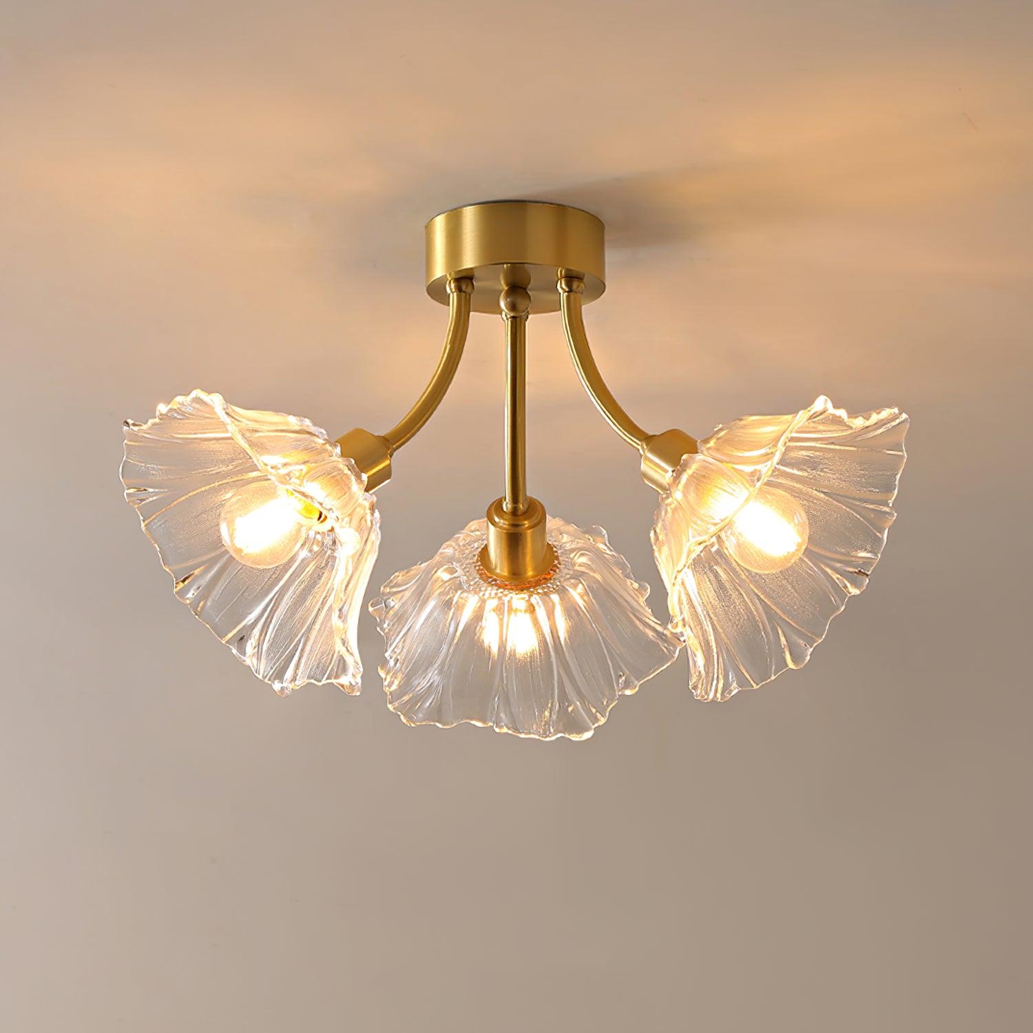 Flush-mount ceiling lamp with curved gold-toned arms holding clear ribbed flower-shaped glass shades, lit, seen from below against a warm wall.