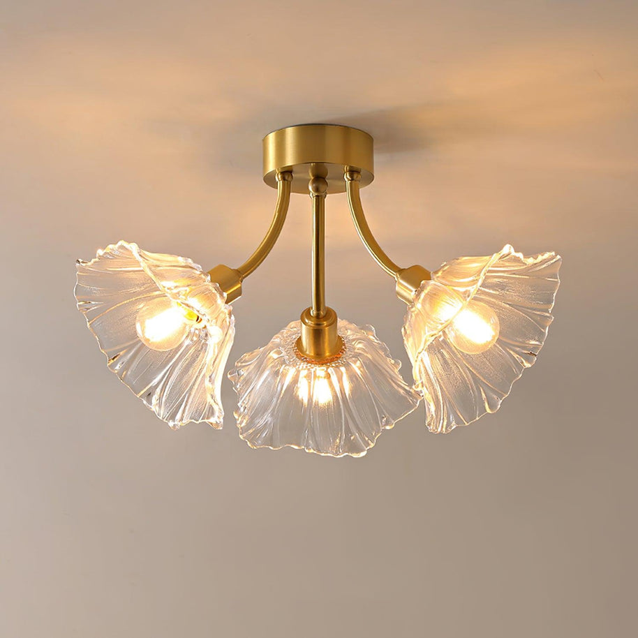 Flush-mount ceiling lamp with curved gold-toned arms holding clear ribbed flower-shaped glass shades, lit, seen from below against a warm wall.