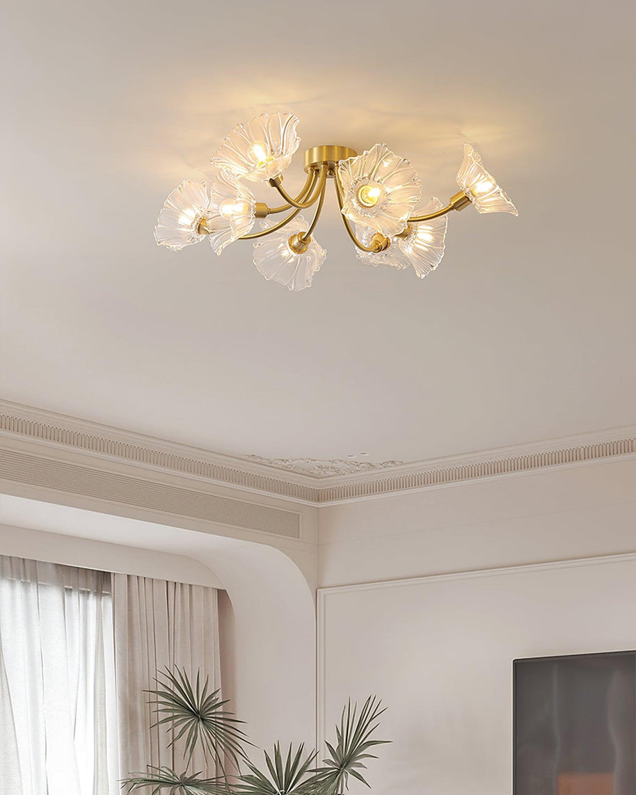 Flush-mount ceiling lamp with curved brass arms holding clear ribbed flower-shaped glass shades lit on a ceiling above a window and a plant.