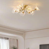 Flush-mount ceiling lamp with curved brass arms holding clear ribbed flower-shaped glass shades lit on a ceiling above a window and a plant.