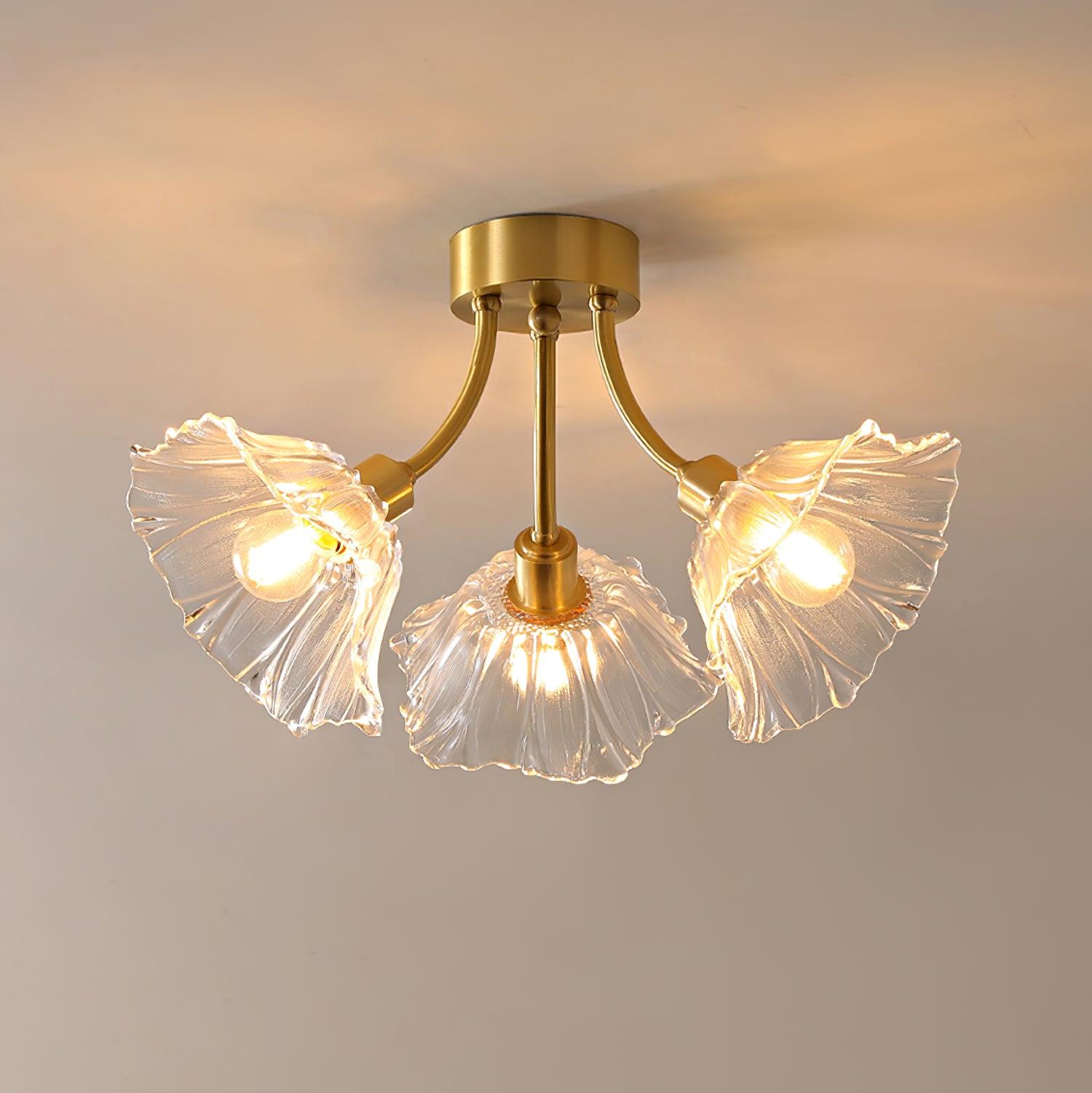 Flush-mount ceiling lamp with curved gold-toned arms holding clear ribbed flower-shaped glass shades, lit, seen from below and slightly to one side.