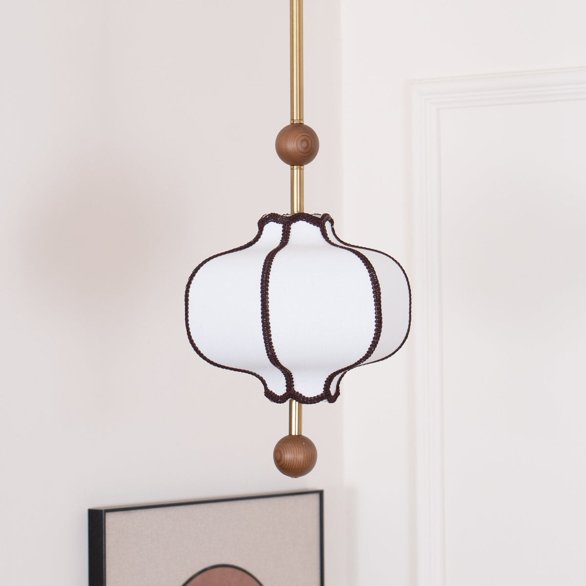 Unlit white fabric lantern pendant with brown braid trim on a segmented brass rod.