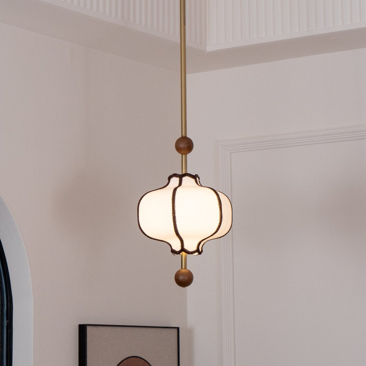 Unlit white lantern pendant hanging on a long brass rod in a white room with a fluted cornice.