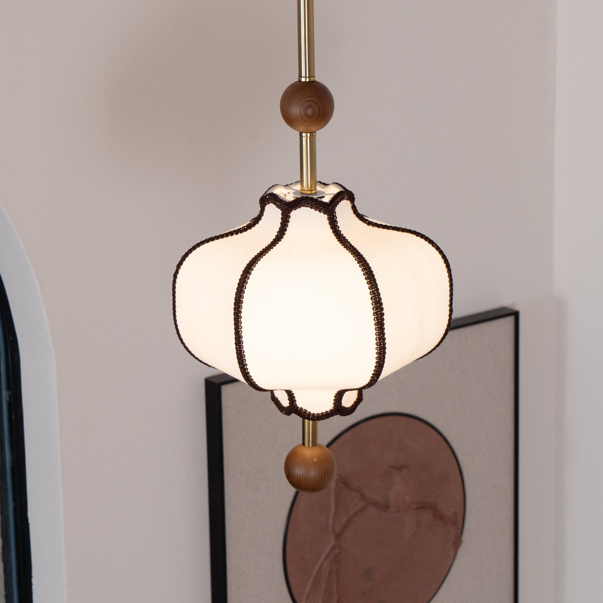 Lit white lantern pendant with brown braid trim beside an arched mirror and a framed print.