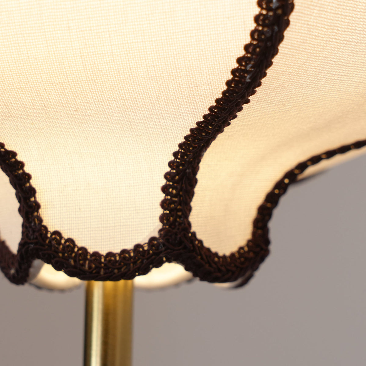 Lit macro of the shade: woven brown braid trim on open-weave white fabric around a brass rod.