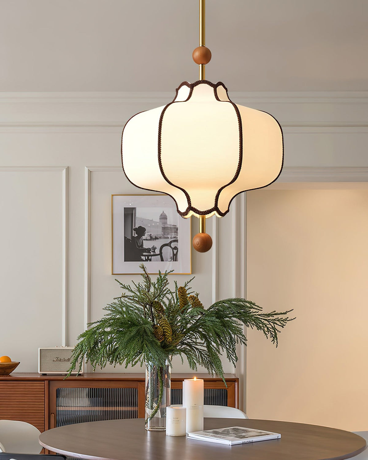 Lit white lantern pendant above a dining table with pine branches and candles in a panelled room.