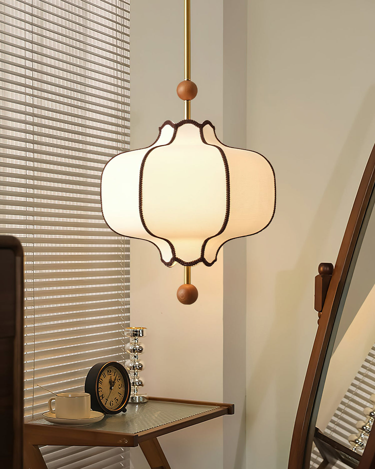 Lit white lantern pendant above a small desk with a clock and a cup, beside white blinds.