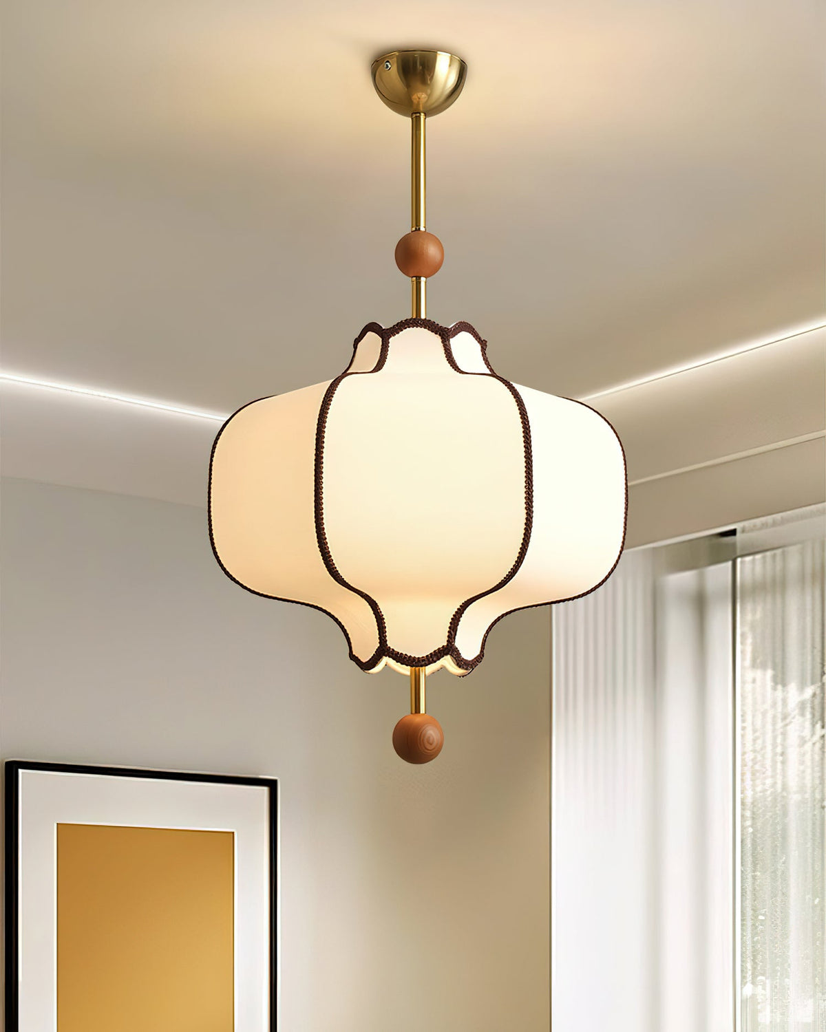Lit white fabric lantern pendant on a brass rod and canopy in a pale room with cove lighting.